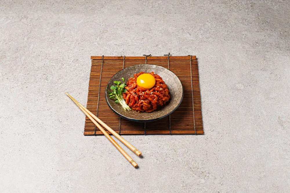 대나무 발 위 검은 접시에 담긴 육회비빔밥을 위에서 비스듬히 촬영한 납품 사진 — 노른자를 올리고 젓가락을 곁들인 구성